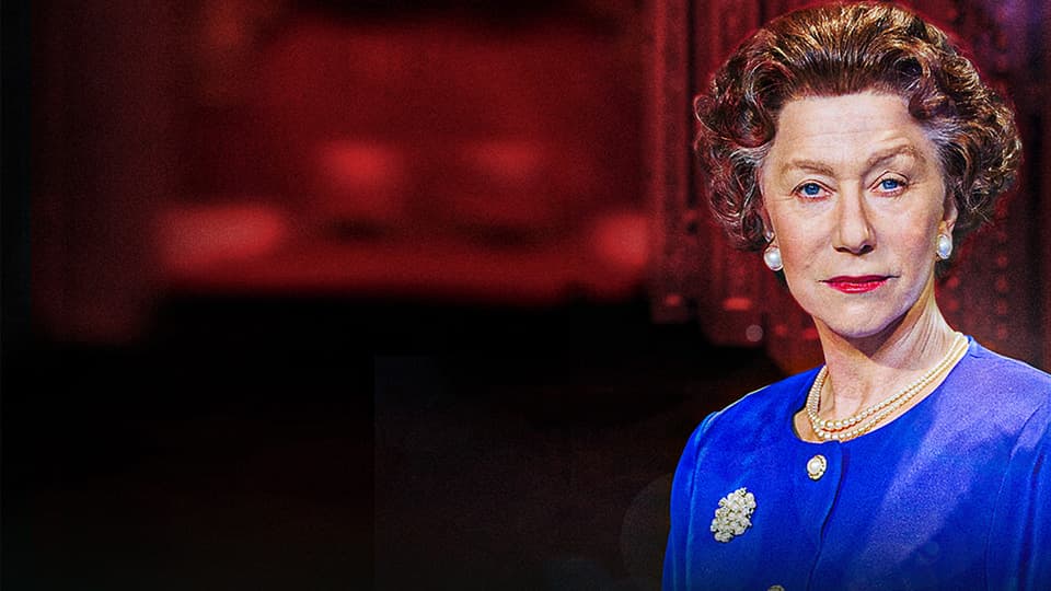 A woman wearing a royal blue suit and pearl necklace, with short brown curly hair, in front of a red background.